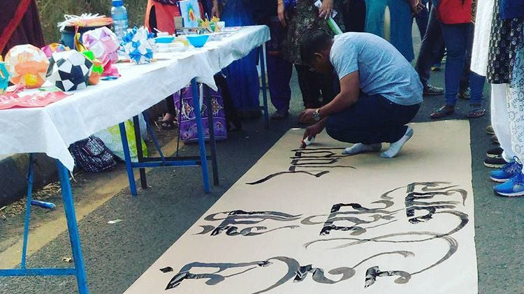 A calligrapher kneels on the floor while drawing some street art as a crowd looks on.