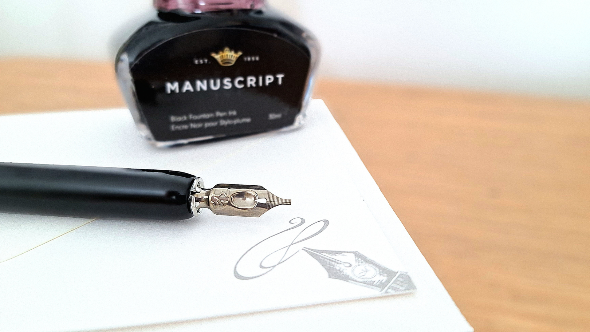 A close up of a dip pen lay on top of manuscript letterhead paper, with a bottle of ink behind.