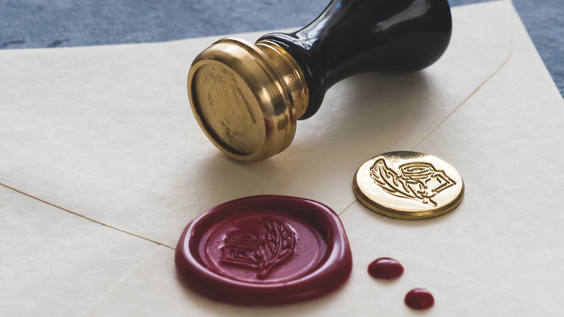 A wax stamp and seal arranged on top of an envelope, the seal is red and shows a quill pen and ink bottle.