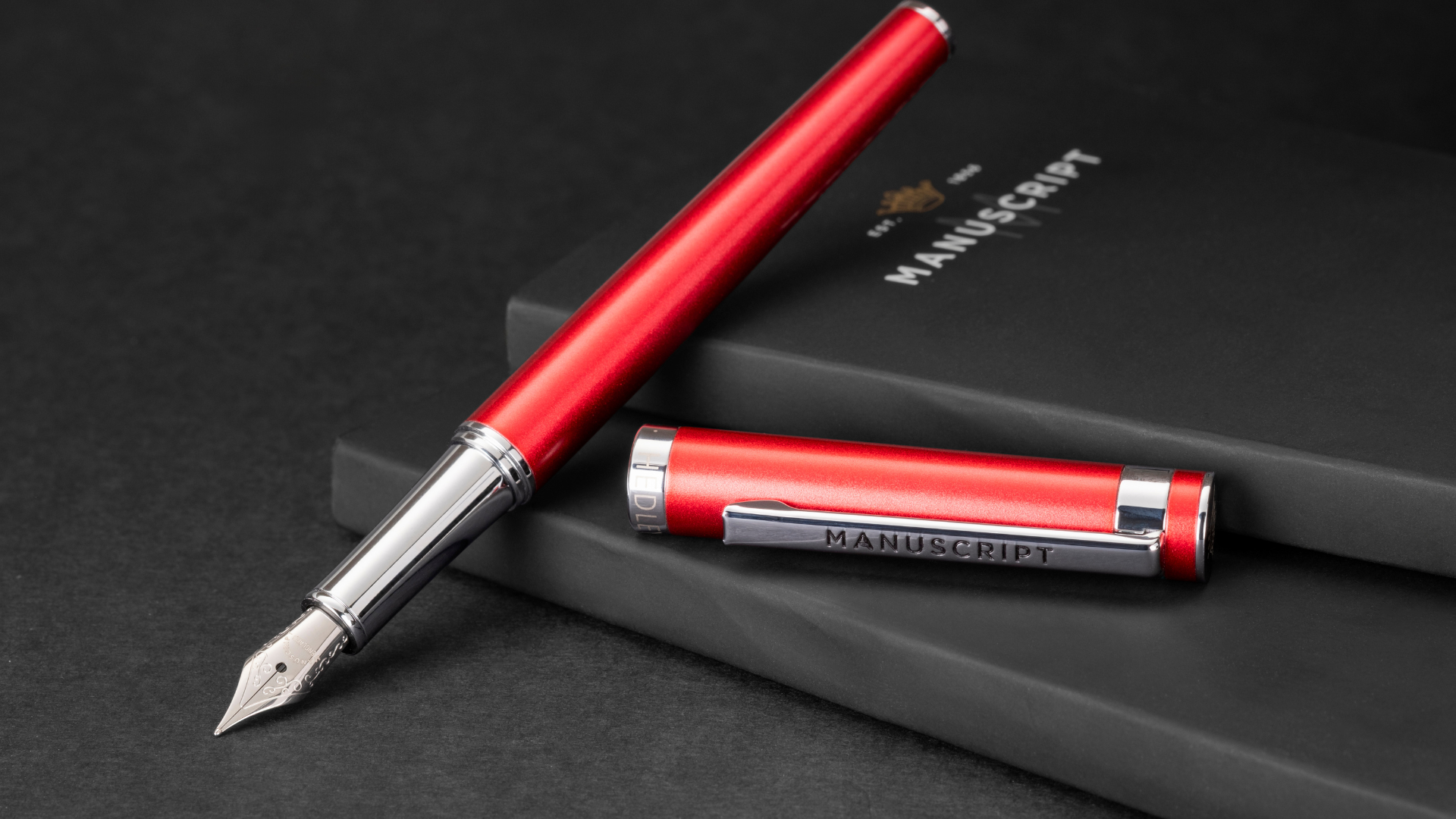 A bright red Hedley range Manuscript fountain pen lays elegantly on top of a black notebook.
