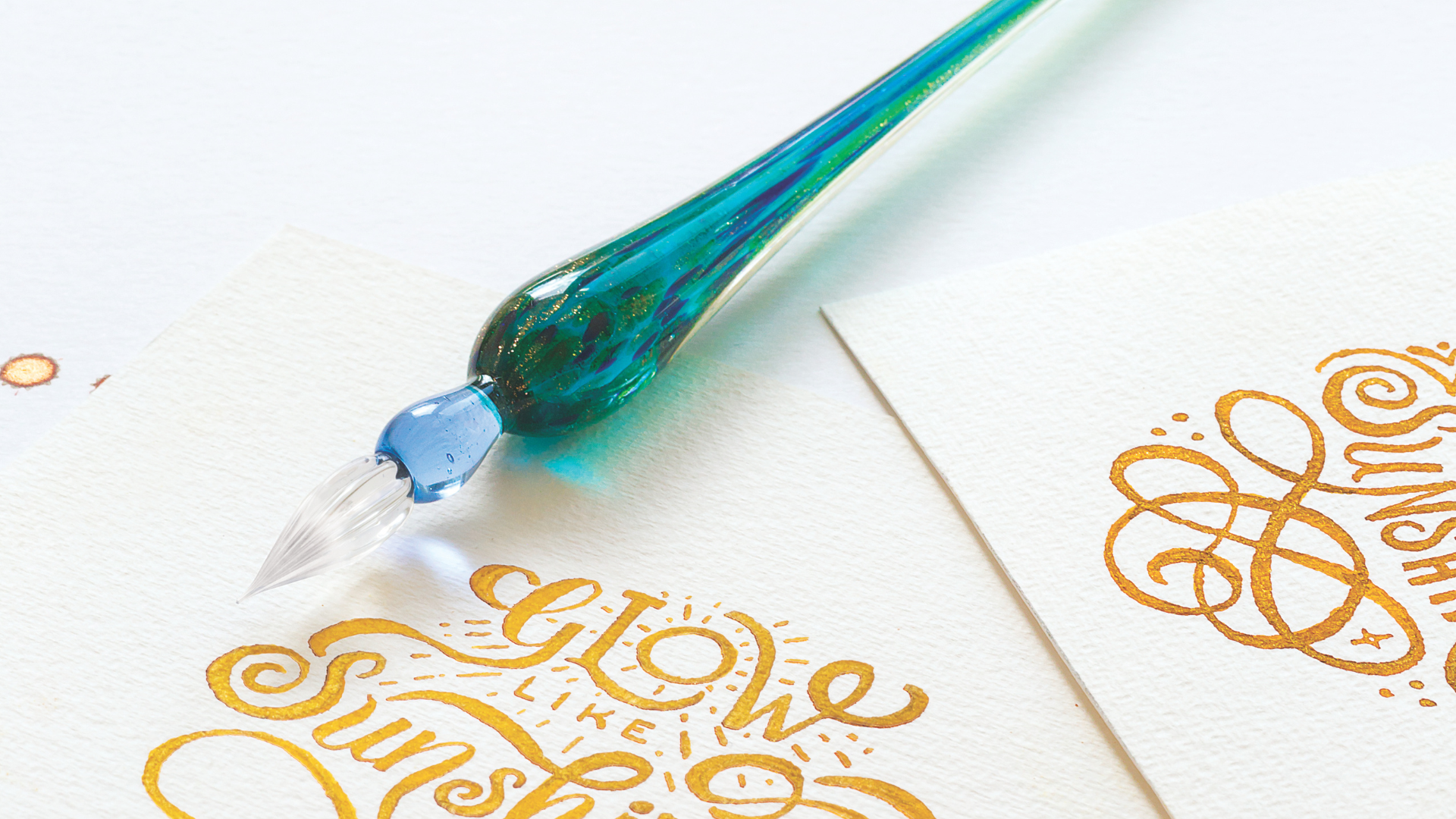 A glass dip pen in turquoise lays on top of a stunning piece of calligaphy, it reads "glow like sunshine", written in gold ink.